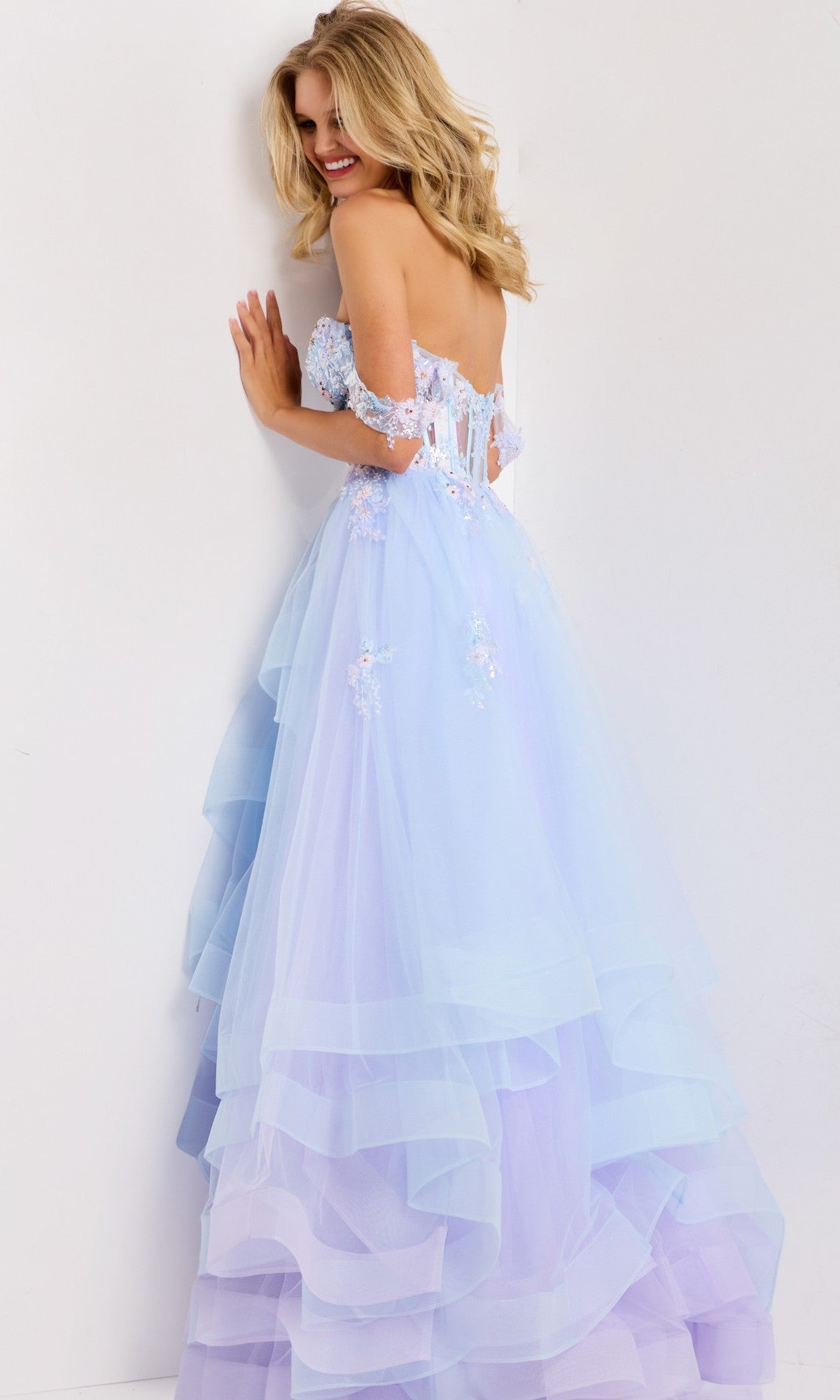 A woman wearing the Jovani 47427 long prom dress, a strapless pastel blue and lavender lilac ballgown with floral appliquรฉs, stands against a white wall, smiling and turning slightly to the side.
